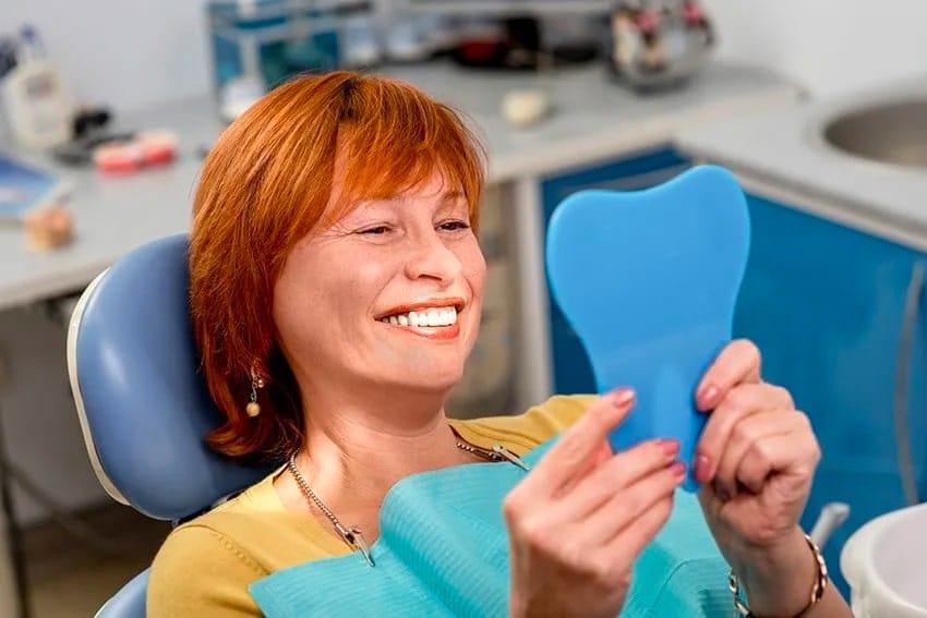 mature red haired woman looks into a handheld mirror at her dentist, smiling