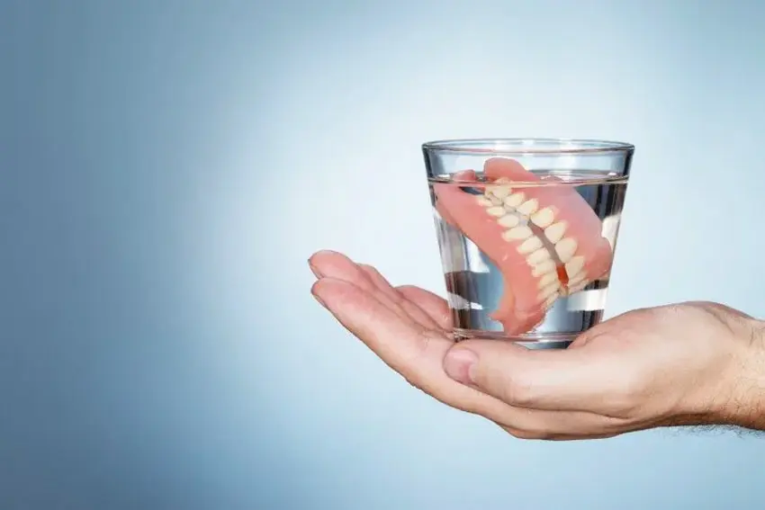 Hand holding up a glass of water with dentures soaking in it