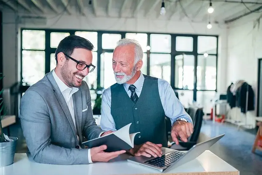 two businessmen, one old, one younger go over a report on a laptop